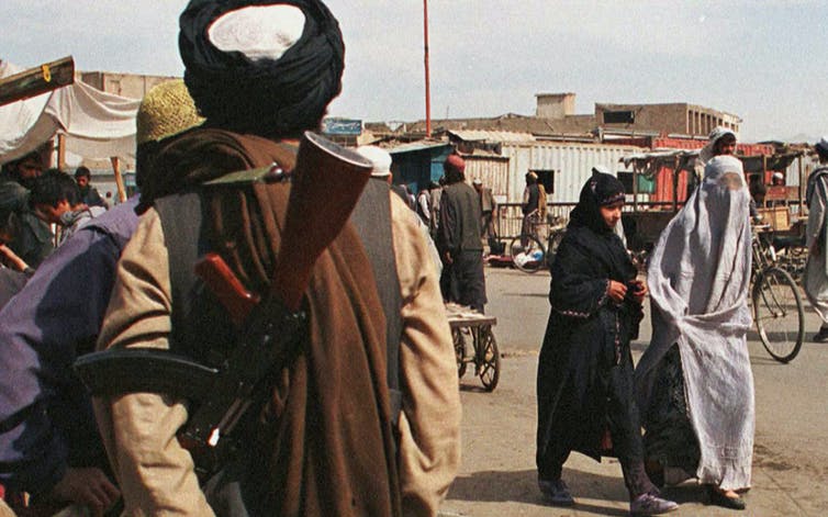 Deux femmes afghanes portant des burqas de styles différents passent devant un soldat de la milice taliban fondamentaliste armé.
