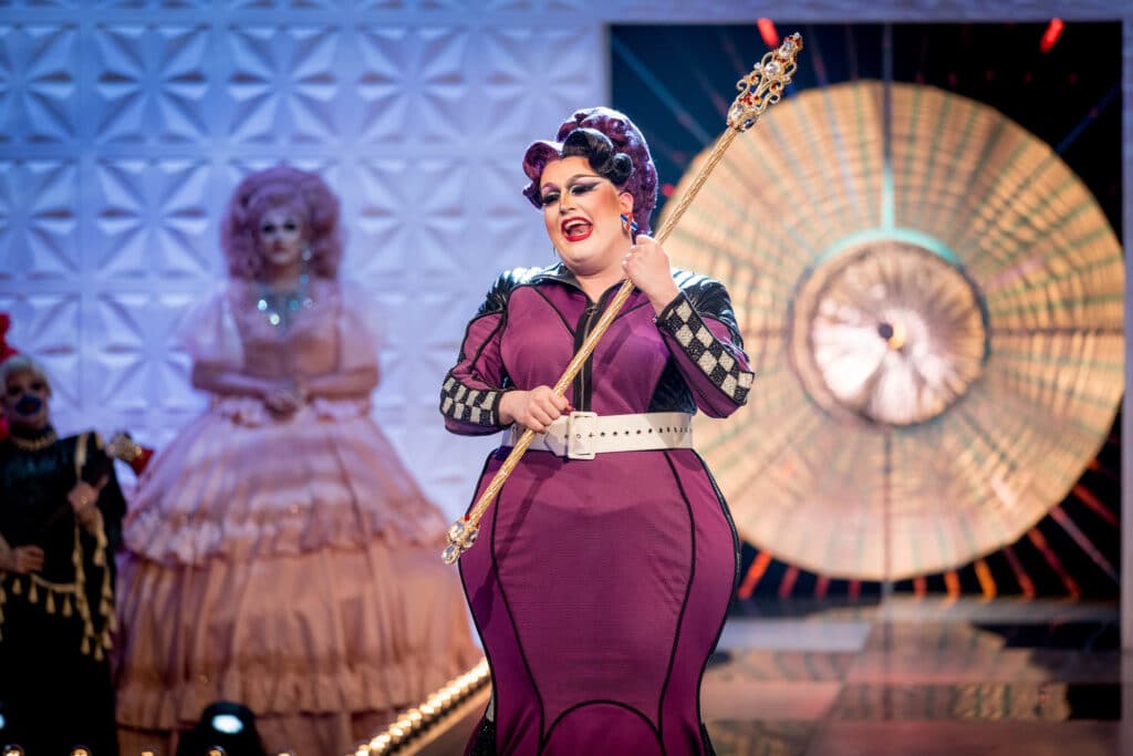 Lawrence Chaney, une drag queen vêtue d'une longue robe plus violette avec un drapeau de course sur les côtés, tenant un sceptre