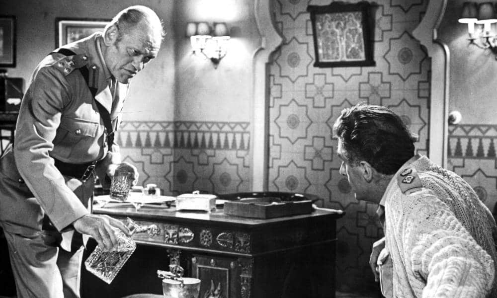 Harry Andrews avec une carafe dans une main et un verre dans l'autre dans une scène du film