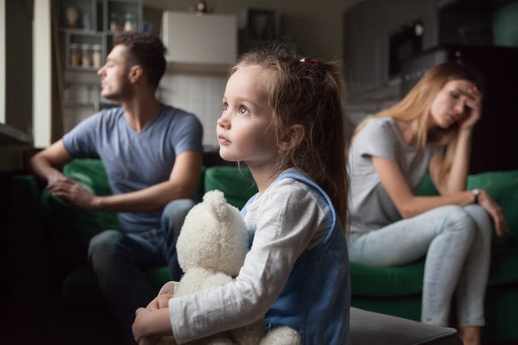 Enfant tenant un ours pendant que les parents ont l'air stressés sur le canapé.