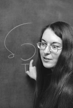 Une femme avec des lunettes pointe vers un tableau.
