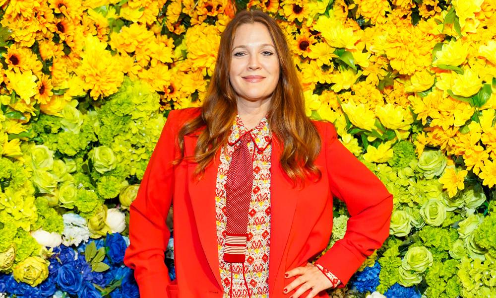 Drew Barrymore porte un haut à motifs et une veste rouge alors qu'elle se tient devant un fond arc-en-ciel composé de fleurs