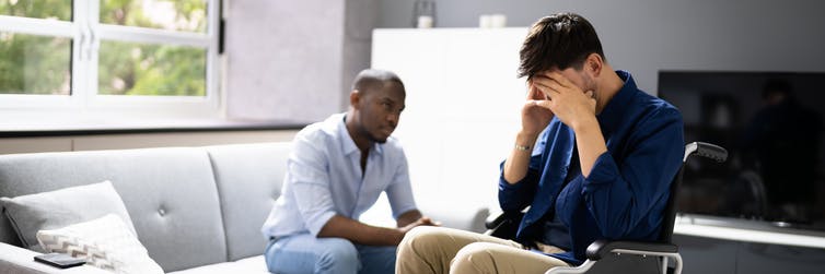 Deux hommes sont assis dans un salon, l'un tenant sa tête dans ses mains.