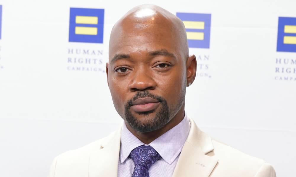 Brian Michael Smith, un acteur trans noir, porte une chemise violette et une cravate violette plus foncée avec une veste de costume blanche