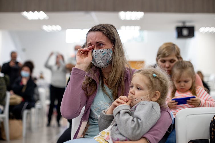 Une femme blonde portant un masque facial tient un enfant dans ses bras et essuie des larmes, dans une pièce d'autres personnes assises.