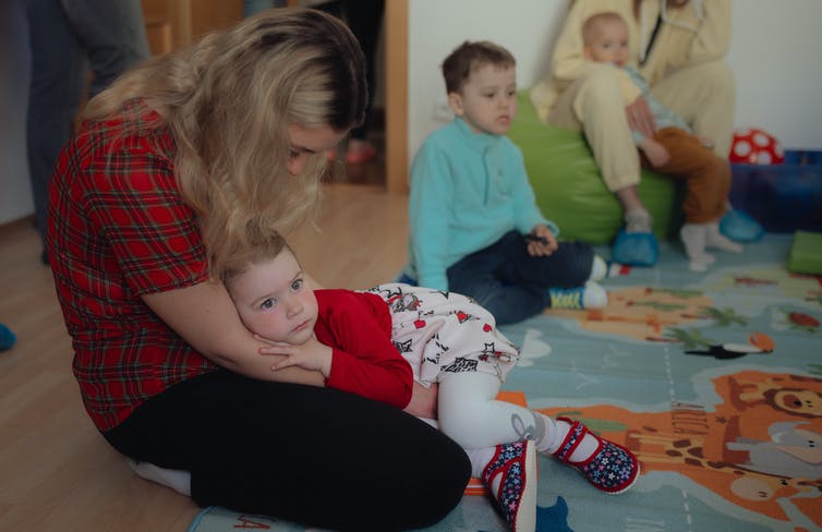 Une femme blonde tient un petit enfant sur ses genoux qui repose contre les bras de sa mère