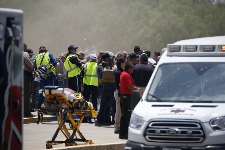 Une foule de personnes en uniformes et gilets de sécurité, debout près d'une ambulance et d'une civière vide.