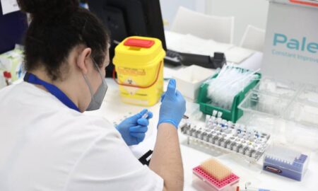 A scientist wearing a grey face mask, blue shirt and white top holds a dropper and container in their blue gloved hands as monkeypox samples are seen on their desk