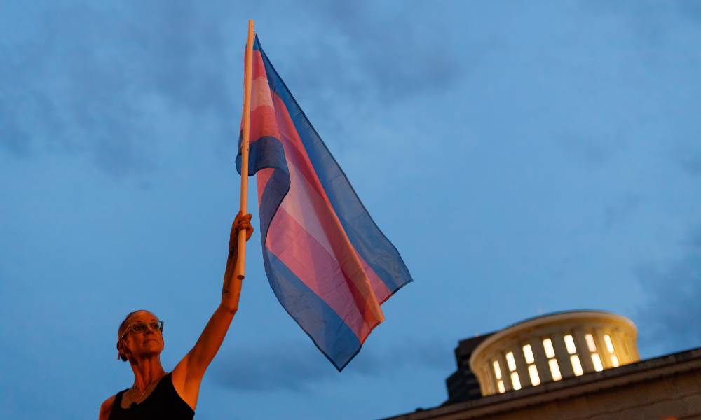 Une personne tient un drapeau transgenre Pride en l'air dans une main devant l'Ohio Statehouse pour protester contre l'adoption d'une interdiction des sports trans