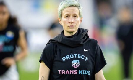 Megan Rapinoe stands on a soccer field while wearing a dark, short-sleeved t-shirt that has the words 'protect trans kids' written on it in blue and pink lettering