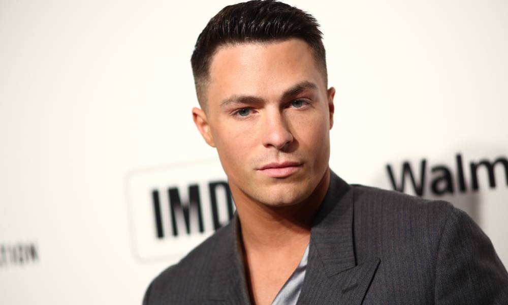 Colton Haynes porte un haut gris avec un morceau de tissu à motifs soyeux sur sa poitrine alors qu'il se tient devant un fond blanc avec le logo IMDb à l'arrière dessus