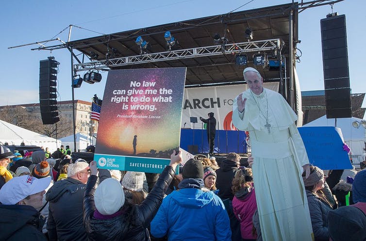 Les participants à un rassemblement tiennent une illustration grandeur nature du pape François devant une scène extérieure.