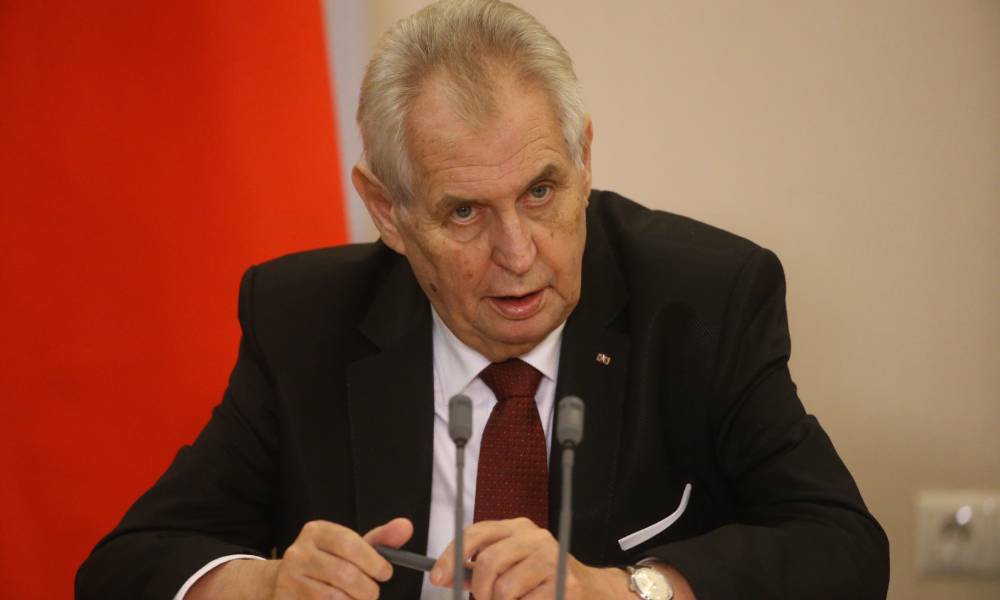 Le président tchèque Miloš Zeman porte une chemise boutonnée blanche, une cravate rouge et une veste noire alors qu'il parle dans deux microphones gris tout en inclinant la tête vers le bas