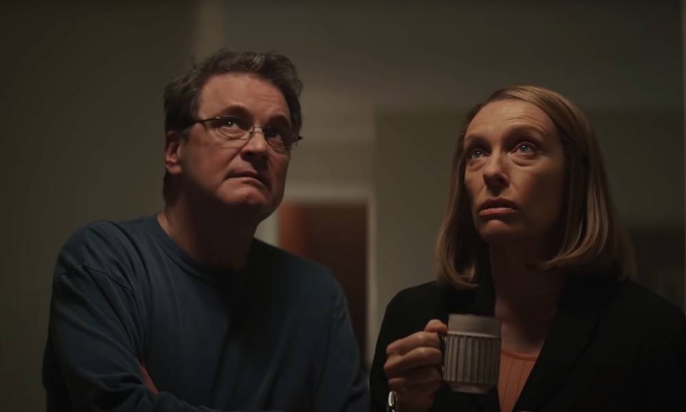 Colin Firth porte un haut sombre et des lunettes alors qu'il joue Michael Peterson dans la série The Staircase de HBO Max.  Toni Collette se tient à côté de lui, portant également un haut sombre et tenant une tasse alors qu'elle représente la femme de Peterson, Kathleen.  Les deux acteurs regardent quelque chose
