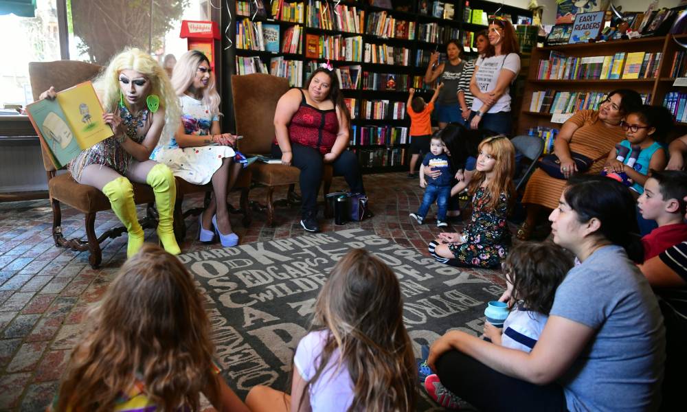 Deux artistes drag sont assis devant une foule d'enfants et d'adultes.  Un interprète lit un livre tandis qu'un autre interprète sourit à la foule