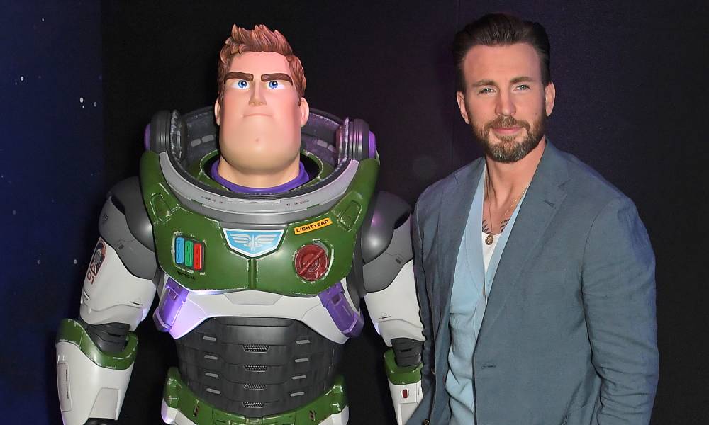 Chris Evans porte une chemise blanche, un pull bleu clair et une veste de costume gris-bleu avec un pantalon assorti alors qu'il se tient à côté d'une statue du personnage de Disney Buzz Lightyear.  Buzz est un personnage qui porte une combinaison spatiale blanche, verte et violette avec un panneau sur la poitrine