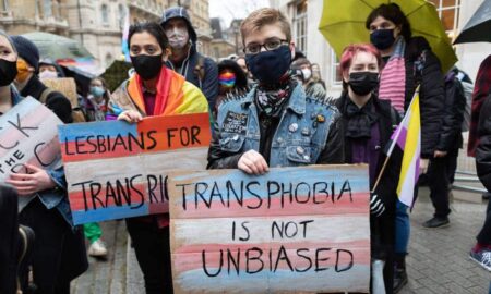 Several people join a protest outside the BBC. One person holds a sign that reads 'Transphobia is not unbiased' that is coloured with blue, pink and white stripes