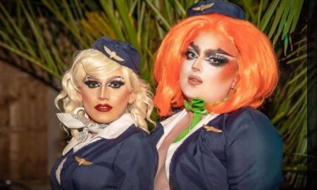 Two drag performers stand side by side, one with blonde hair and another with orange hair, as they appear to wear what looks like air cabin crew uniforms