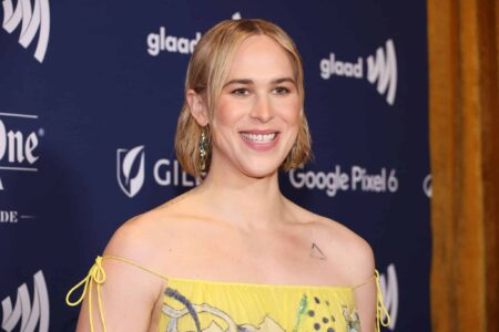 Tommy Dorfman stands in the press section of the GLAAD Media Awards wearing a yellow dress.