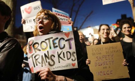 A person in gather amid a crowd in a protest holding a sign that reads 'Protect trans kids'