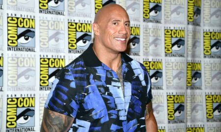 Dwayne 'The Rock' Johnson smiles for the camera as he wears a blue and black patterned top and stands in front of a patterned background with the SD Comic Con logo on it