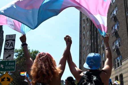 In this photograph, two trans people hold hands while marching under a trans pride flag