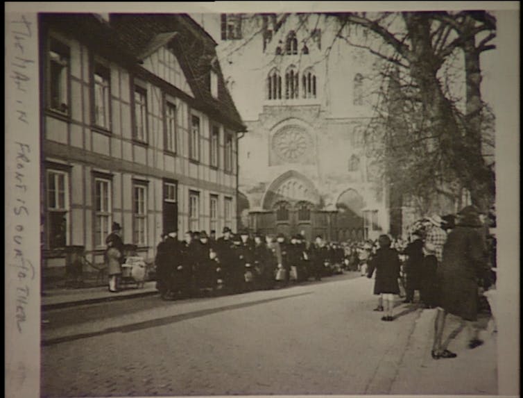 Un grand groupe de personnes s'est rassemblé dans la rue devant un bâtiment à colombages et une grande église, avec des gens qui les regardaient de l'autre côté de la rue.