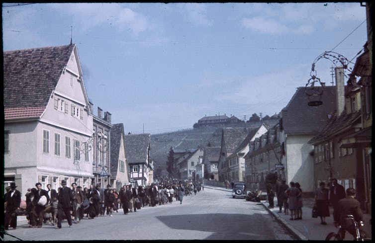 Des centaines de personnes défilent dans une rue du village, tandis que les spectateurs regardent.