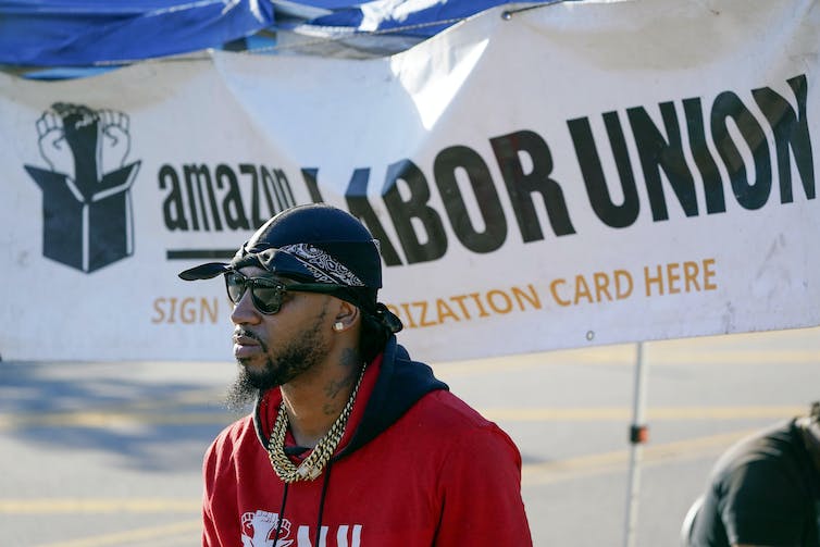 Un homme portant un sweat-shirt rouge se tient devant une bannière « Amazon Labour Union ».