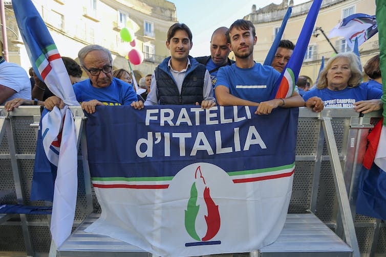 Un groupe d'hommes se tient autour d'un drapeau avec 'Fratelli d'Italia' dessus et un symbole de flamme en dessous.