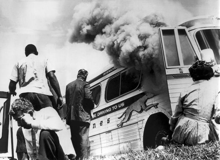 Photographie en noir et blanc de personnes debout et assises à l'extérieur d'un autobus en feu.