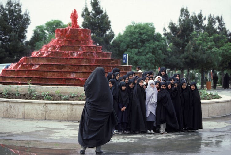Un groupe de filles en robe noire pose devant une fontaine pendant qu'une femme prend leur photo.