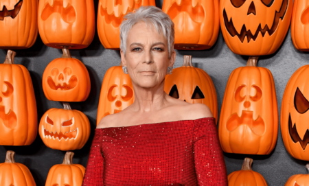 Jamie Lee Curtis wears a red outfit as she stands in front of a wall filled with orange pumpkin items shaped to look like jack-o-lanterns