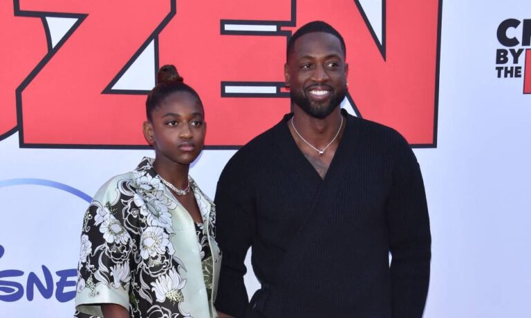 Dwyane Wade wears a black shirt and smiles as he stands next to his daughter Zaya who is wearing a black and white floral patterned top
