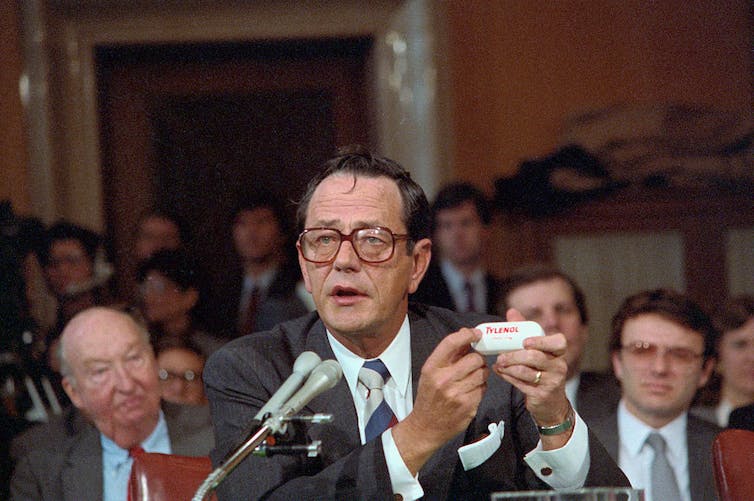 Un homme portant des lunettes et un costume parle dans le microphone tout en tenant une bouteille blanche de pilules.