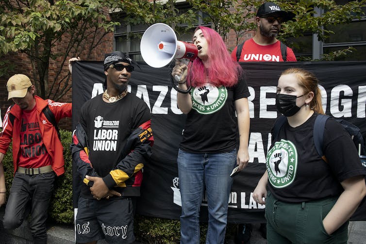 Une femme aux cheveux roses parle dans un mégaphone entourée d'autres organisateurs syndicaux vêtus de noir