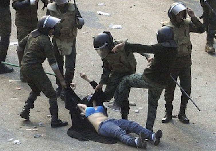 Une femme à moitié vêtue portant un soutien-gorge bleu et un jean traînée par des policiers, l'un d'eux étant sur le point de lui marcher dessus.