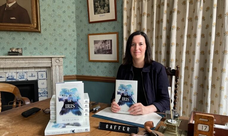 Author Beth Lewis wearing a black top and jacket sits behind a desk holding a copy of her book The Origins of Iris.