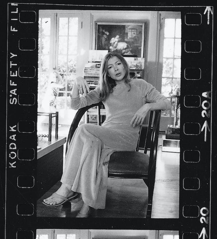Portrait noir et blanc.  Joan est assise chez elle dans une robe à la cheville, regardant la caméra, une jambe croisée sur l'autre.