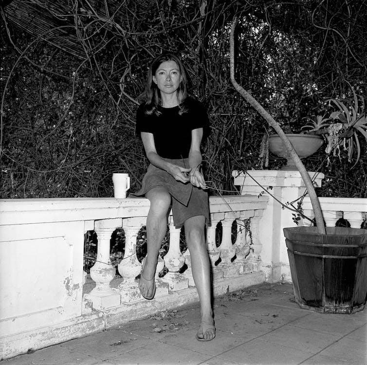 Portrait noir et blanc.  Joan Didion est assise sur un muret dans une jupe jusqu'aux genoux, les cheveux longs sur les épaules.  Une tasse est perchée à sa gauche.