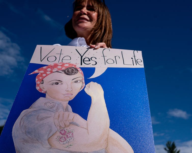 Une femme blanche aux cheveux bruns brandit une affiche Rosie the Riveter qui dit 