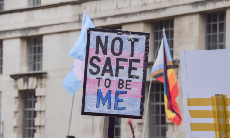 A person holds up a sign, designed in the colours of the trans Pride flag, that reads 'not safe to be me'