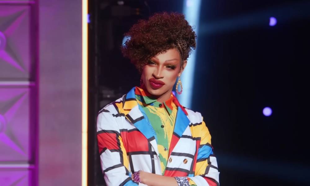 La star de Drag Race, Yvie Oddly, porte une couche extérieure à motifs géométriques blancs, bleus, rouges et jaunes avec une autre couche colorée de vêtements en dessous.  Les cheveux d'Yvie sont coiffés en chignon