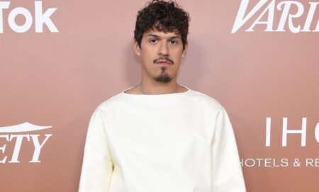 Omar Apollo wears a white top as he stares at the camera while standing in front of a rose pink background