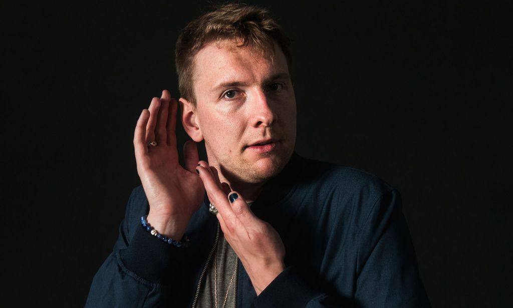 Joe Lycett pose avec ses mains près de son visage alors qu'il regarde vers la caméra