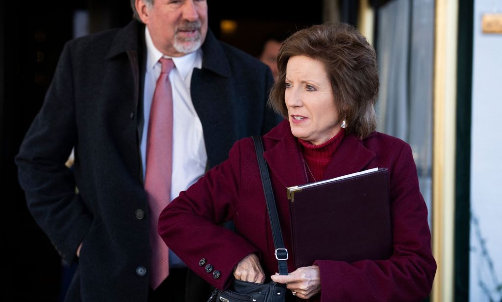 Vicky Hartzler, vêtue d'un manteau rouge foncé, fouille dans son sac tout en regardant vers la droite, un grand homme plus âgé se tient derrière elle avec une cravate rose.