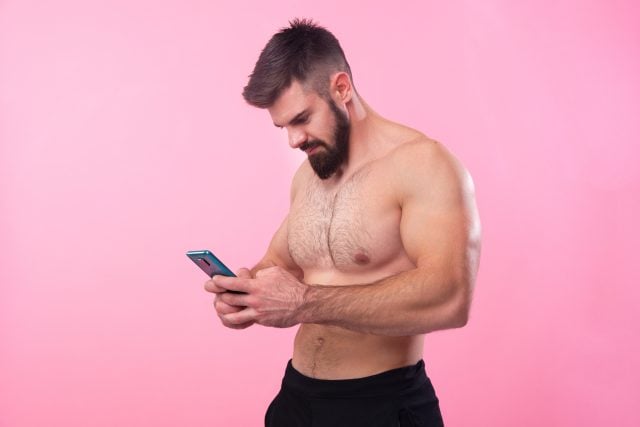 Bodybuilder fort torse nu parlant sur sa cellule, à l'aide d'un smartphone, fond de studio rose