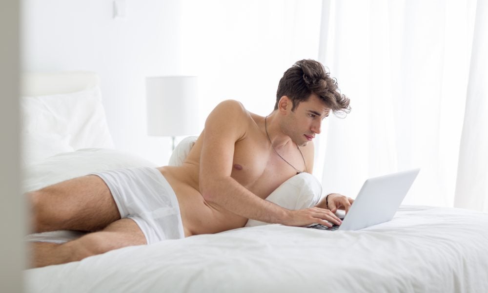 Grindr Desktop : Comment utiliser Grindr sans l’application Young shirtless man working on a laptop computer lying in bed during the day