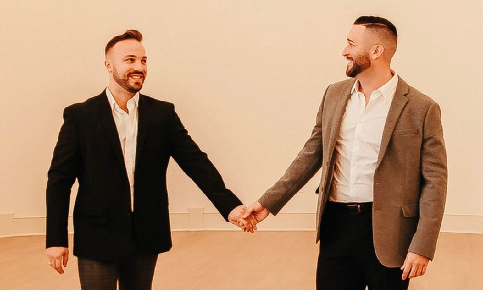 La star du baseball TJ House se révèle fièrement gay et annonce ses fiançailles avec son petit ami : « Je suis aimé » TJ House and Ryan Neitzel hold hands while looking at one another in an empty white room with wall lights above them.