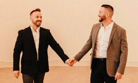 TJ House and Ryan Neitzel hold hands while looking at one another in an empty white room with wall lights above them.